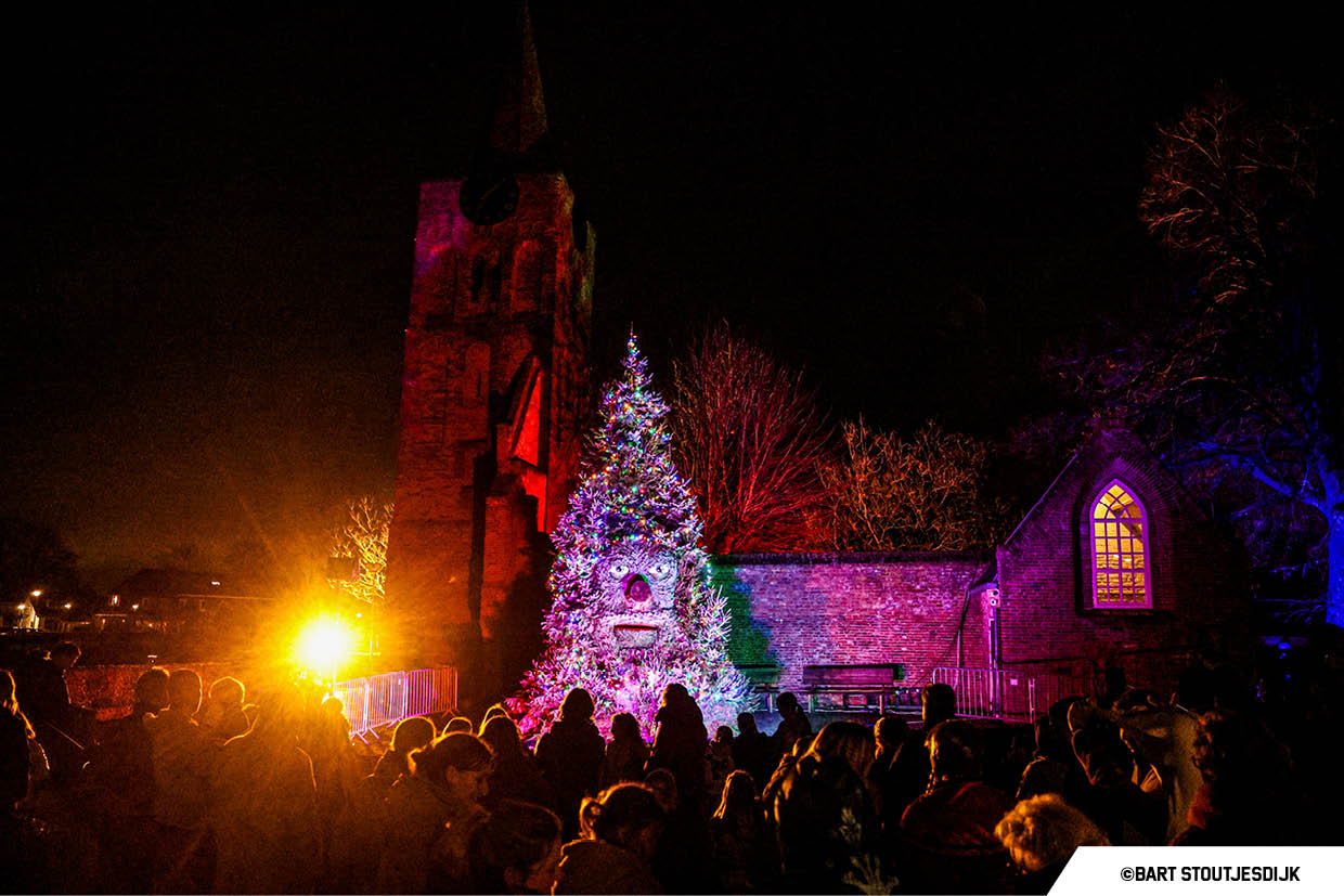 Publiek bekijkt een verlichte kerstboom voor een kerk op een winteravond