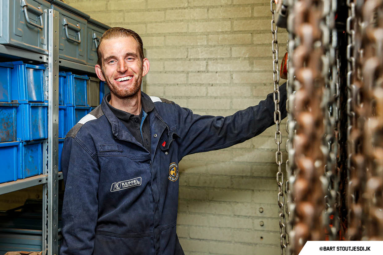 Glimlachende medewerker in overall van Keppel Constructie staat in een werkplaats tussen stellingen en hijskettingen