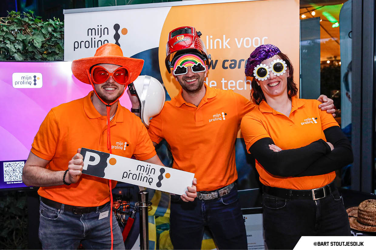 Drie medewerkers in oranje polo's poseren met feestelijke accessoires bij de stand van Prolinq op Werkend Altena