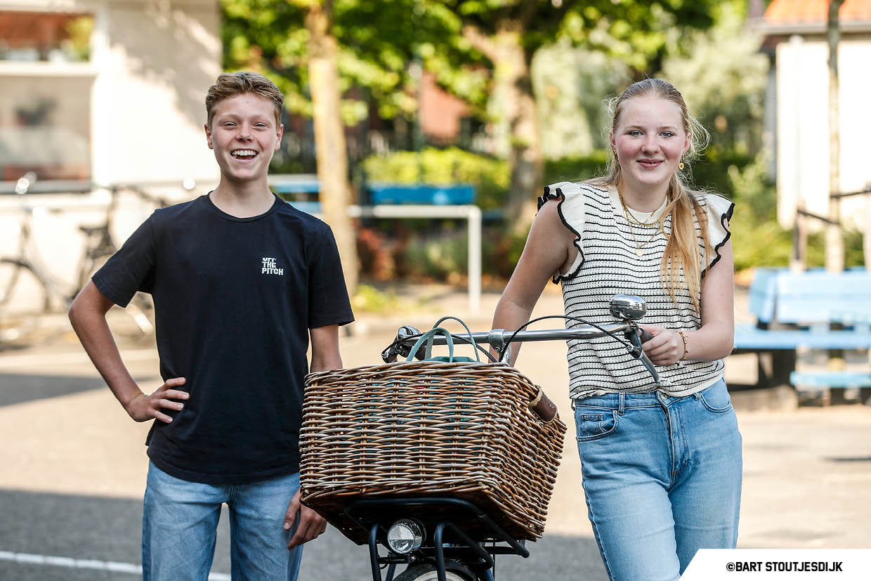 Twee leerlingen poseren glimlachend bij een transportfiets met grote rieten mand