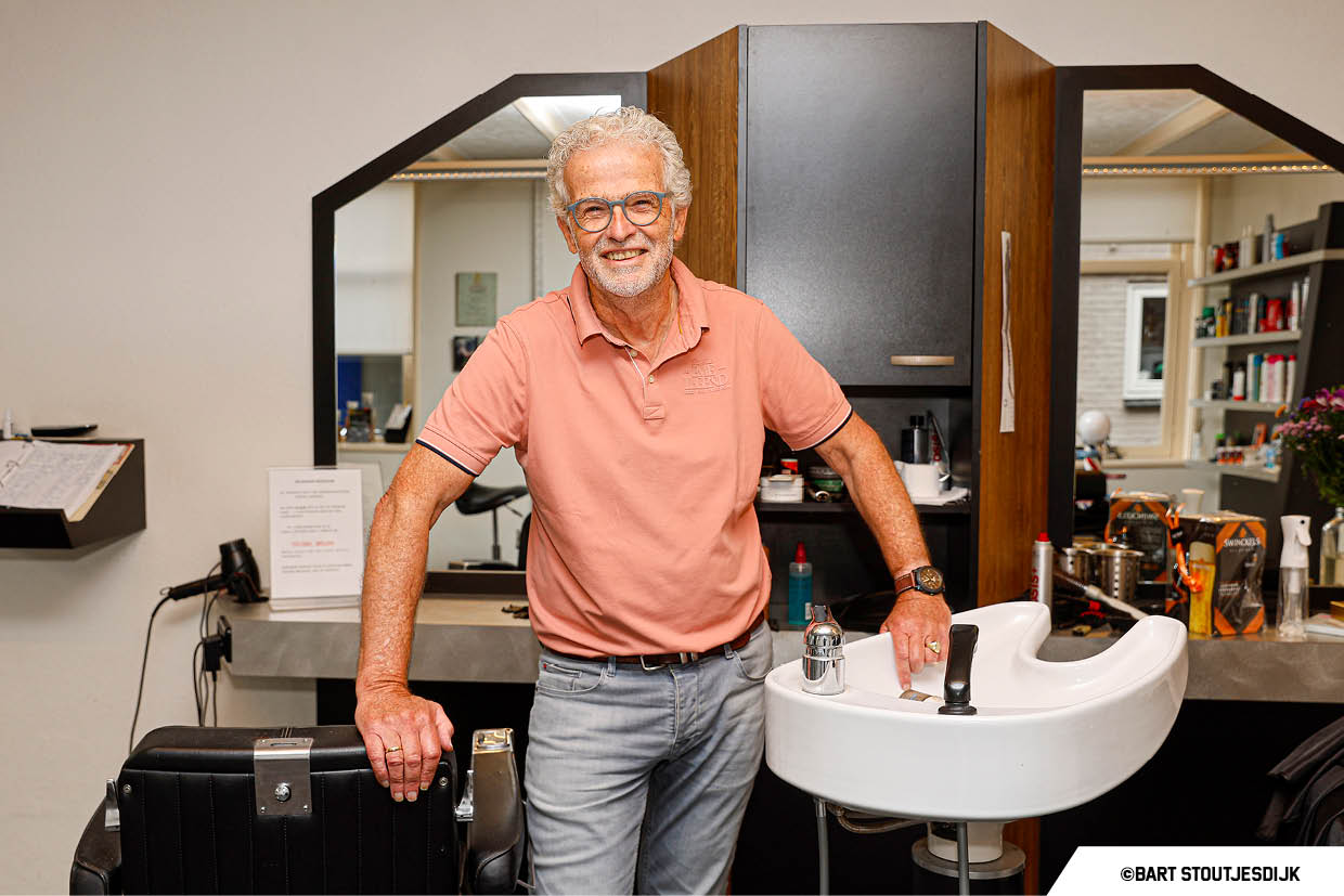 Kapper glimlacht in zijn salon tussen spiegel, wasbak en productplanken