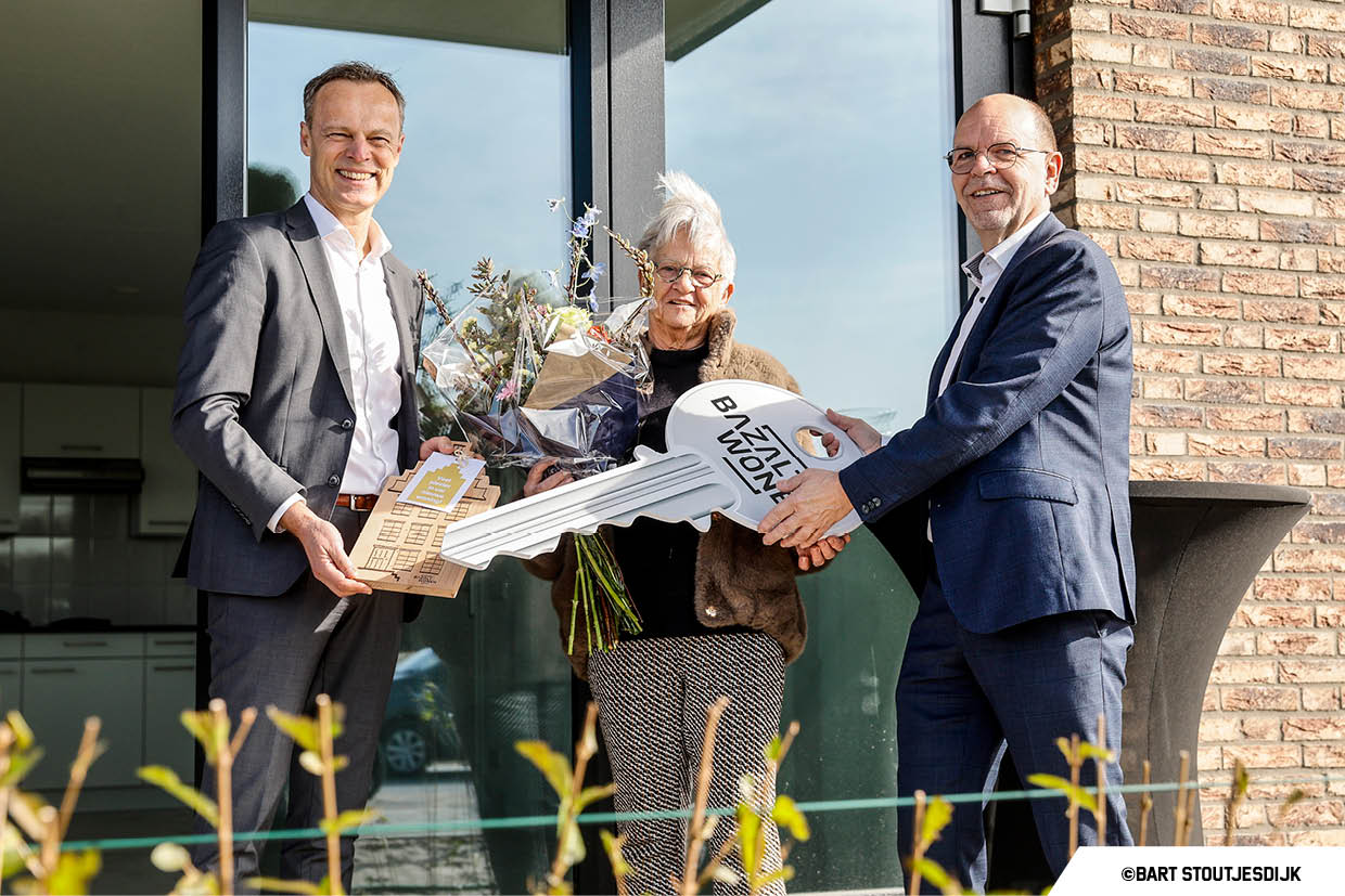 Een bewoonster ontvangt een grote witte sleutel en een houten plaque bij een nieuw huis van Bazalt Wonen
